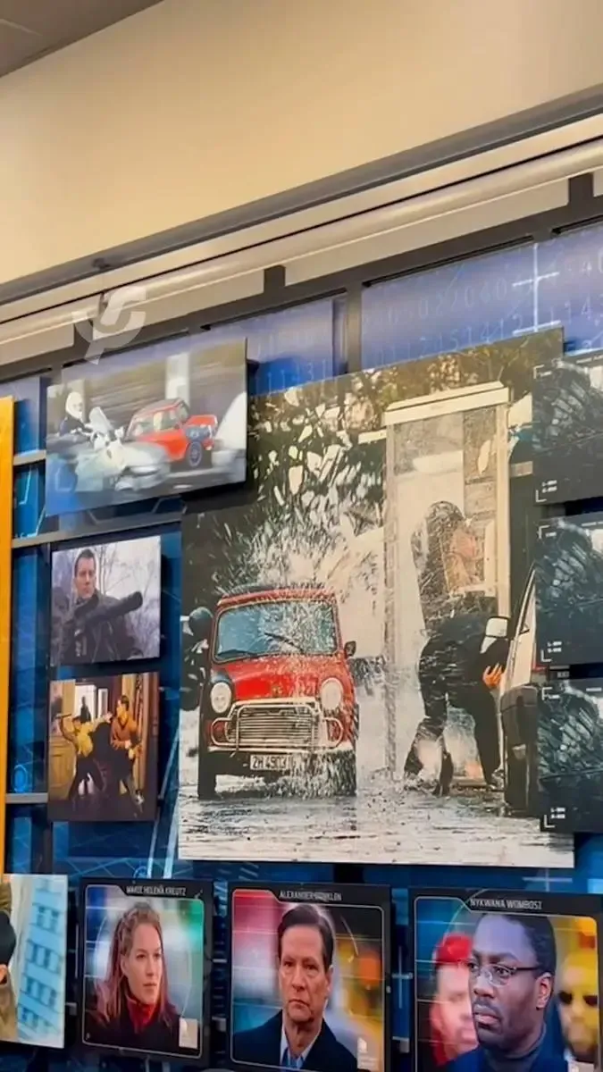 A wall display features various action movie scenes, including a prominent image of a red Mini Cooper splashing through water near a man exiting a car. Below, there are smaller photos of intense action moments and three framed portraits of individuals with serious expressions.