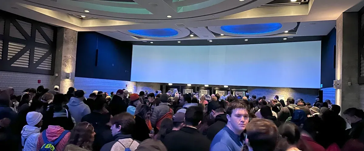 A large crowd of people fills a spacious indoor area with a modern ceiling featuring blue circular lights. Many are wearing jackets and hats, suggesting a cold environment outside. The atmosphere is bustling, with individuals facing various directions, possibly waiting in line.