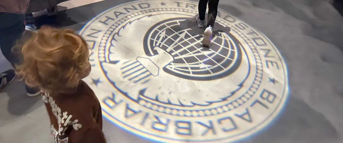 A child with curly hair stands near a large, projected emblem on the floor, featuring a globe and the words "BLACKBIRD" and "TREADSTONE." Another person is walking across the emblem, adding a sense of motion to the scene.