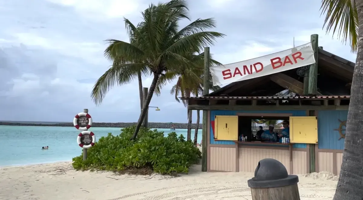 A beachside bar labeled "Sand Bar" is nestled among palm trees, with a vibrant blue ocean in the background. The bar has a rustic appearance with an open window where people are being served. A lifebuoy sign and lush greenery add to the tropical setting.
