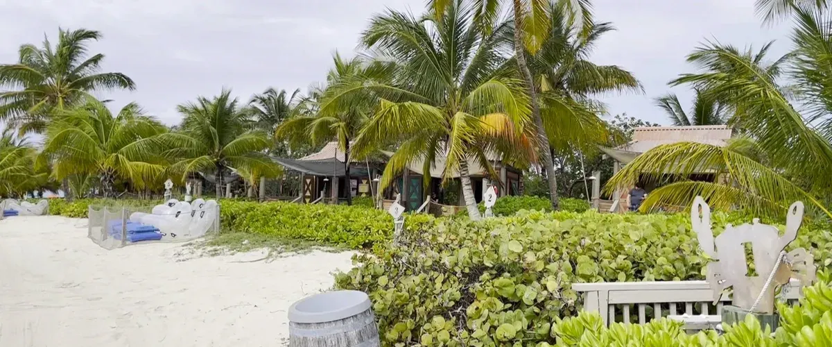 A sandy beach with lush green palm trees and bushes in the foreground, leading to a small resort with thatched-roof buildings. Blue lounge chairs are visible on the sand, and the sky is overcast, creating a tranquil, tropical atmosphere.