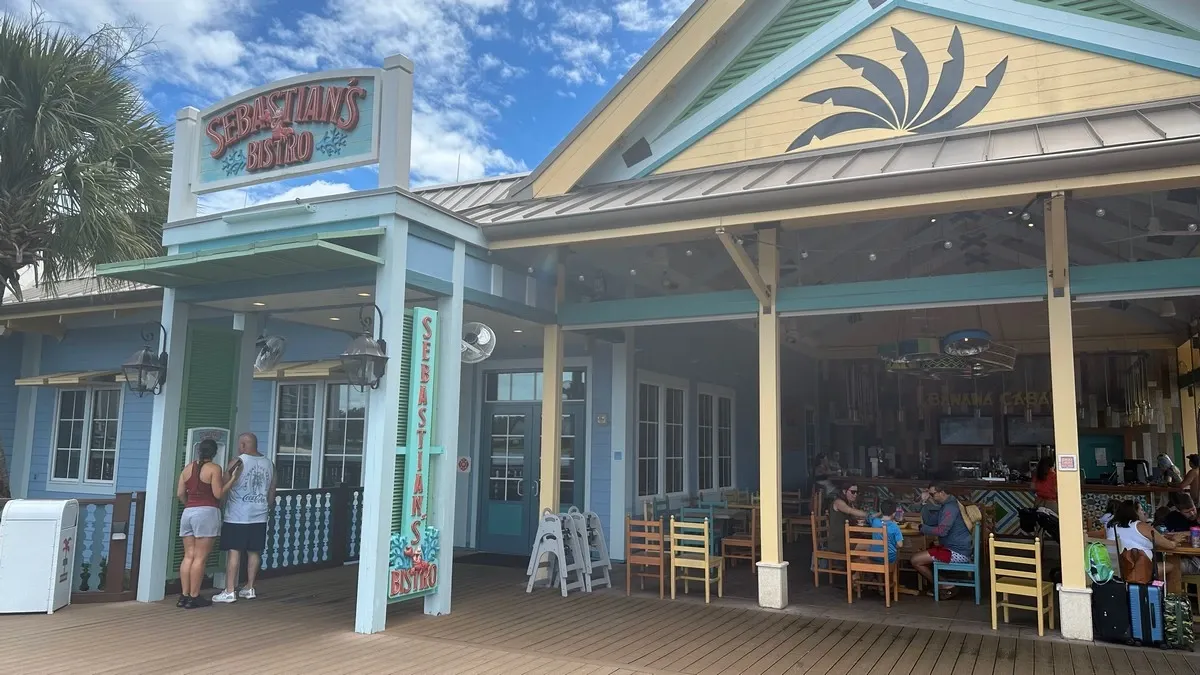 A colorful seaside restaurant with a sign reading "Sebastian's Bistro" featuring an outdoor seating area with wooden chairs and tables. The building is painted in pastel shades with decorative elements, set against a backdrop of a clear blue sky.