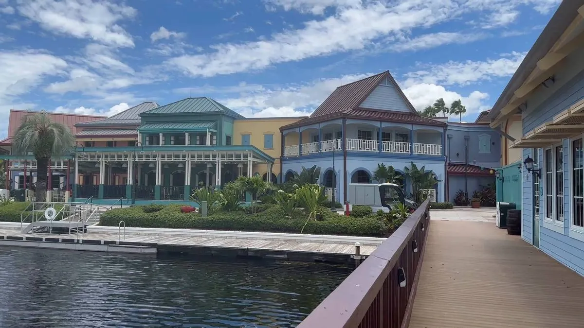 Colorful Caribbean-style buildings line a waterfront walkway under a vibrant blue sky, with greenery and a calm body of water creating a serene atmosphere.