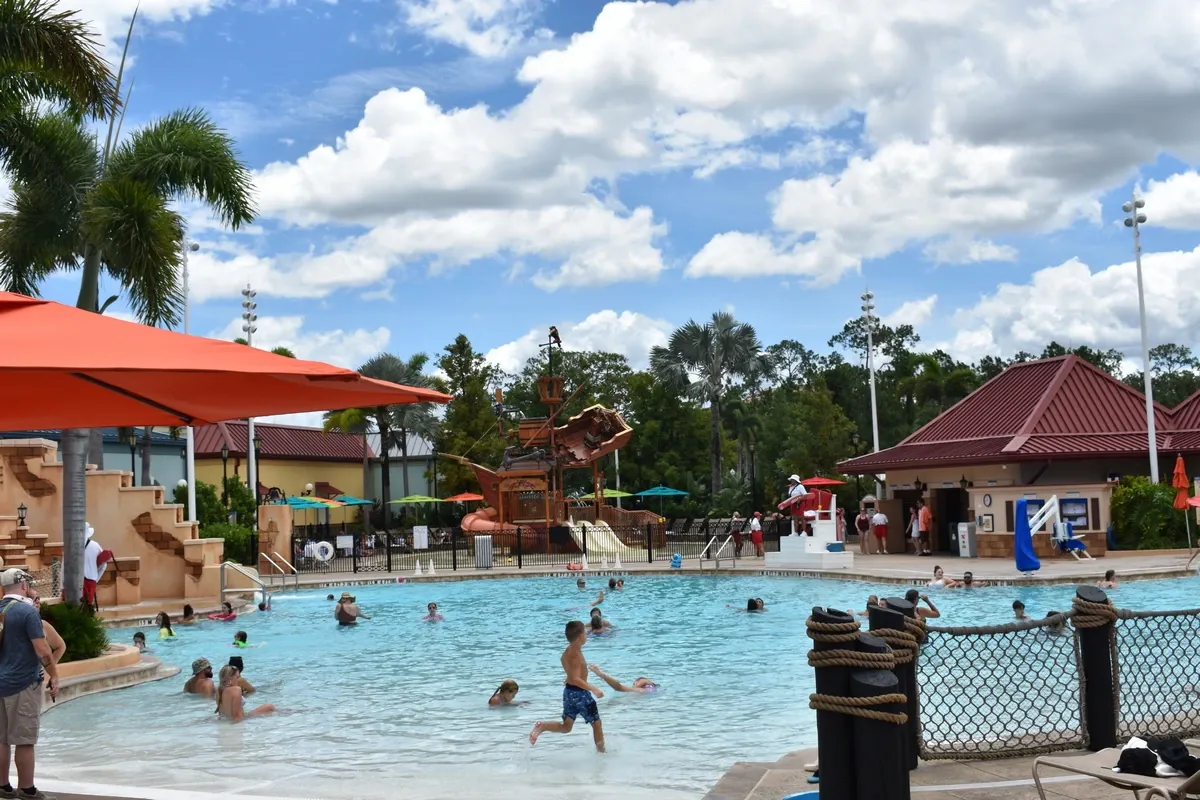 A swimming pool scene featuring people enjoying the water under a partly cloudy sky. An orange umbrella provides shade near the pool, surrounded by palm trees and a waterslide shaped like a ship.