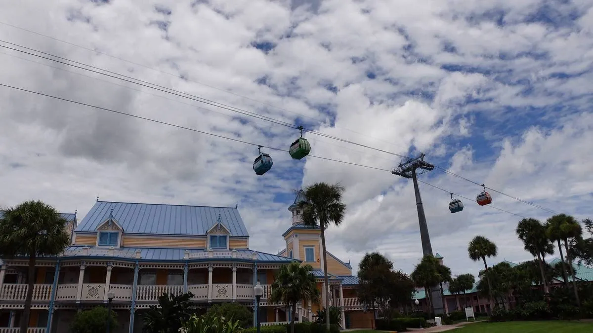 Colorful gondolas glide along a cable above a pastel-colored building with a blue roof, set against a partly cloudy sky with palm trees scattered around.