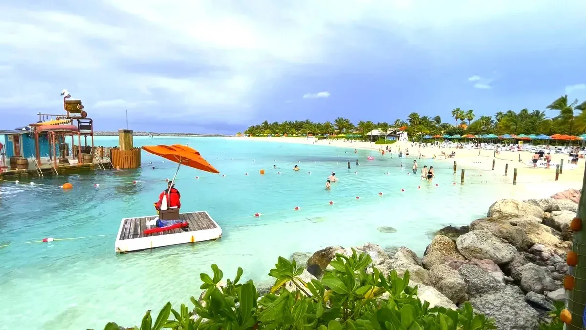 A vibrant beach scene features a lifeguard sitting under an orange umbrella on a floating platform in the turquoise water, with people swimming and relaxing on the sandy shore lined with palm trees.