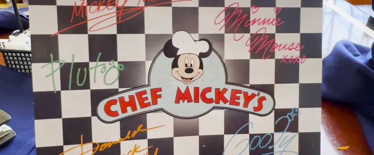 A checkered Chef Mickey's menu featuring colorful autographs of Disney characters, including Mickey Mouse, Minnie Mouse, Pluto, Donald Duck, and Goofy, with a cheerful Mickey illustration in a chef's hat at the center.