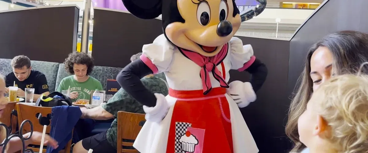Minnie Mouse, dressed in a red and white outfit with a cupcake design, interacts with a young child at a restaurant. In the background, people are seated at tables, enjoying their meals.