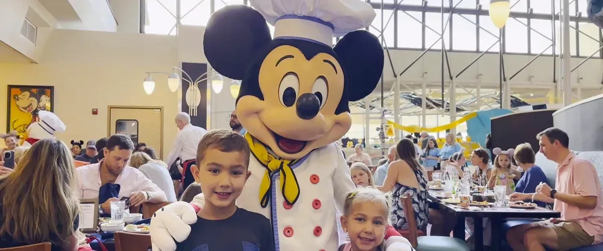 A person in a Mickey Mouse costume dressed as a chef poses with two smiling children in a bustling restaurant filled with families and diners.