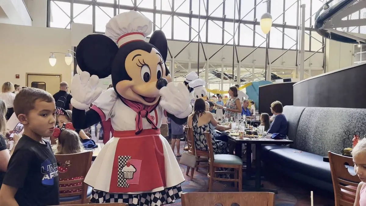 Minnie Mouse, dressed as a chef with a red and white outfit, entertains guests at a lively restaurant filled with families and children. 