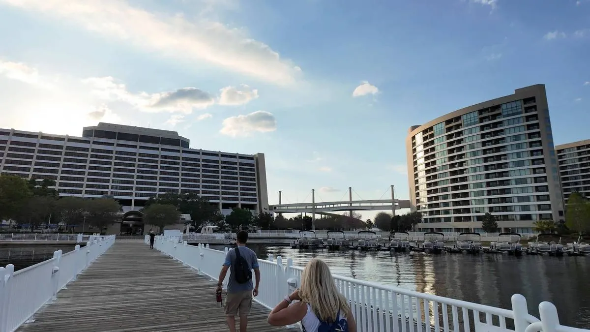 Two people walk along a wooden pier bordered by white railings, leading towards a large hotel with modern architecture. The scene is set by a calm body of water reflecting the buildings, with a blue sky and scattered clouds overhead.