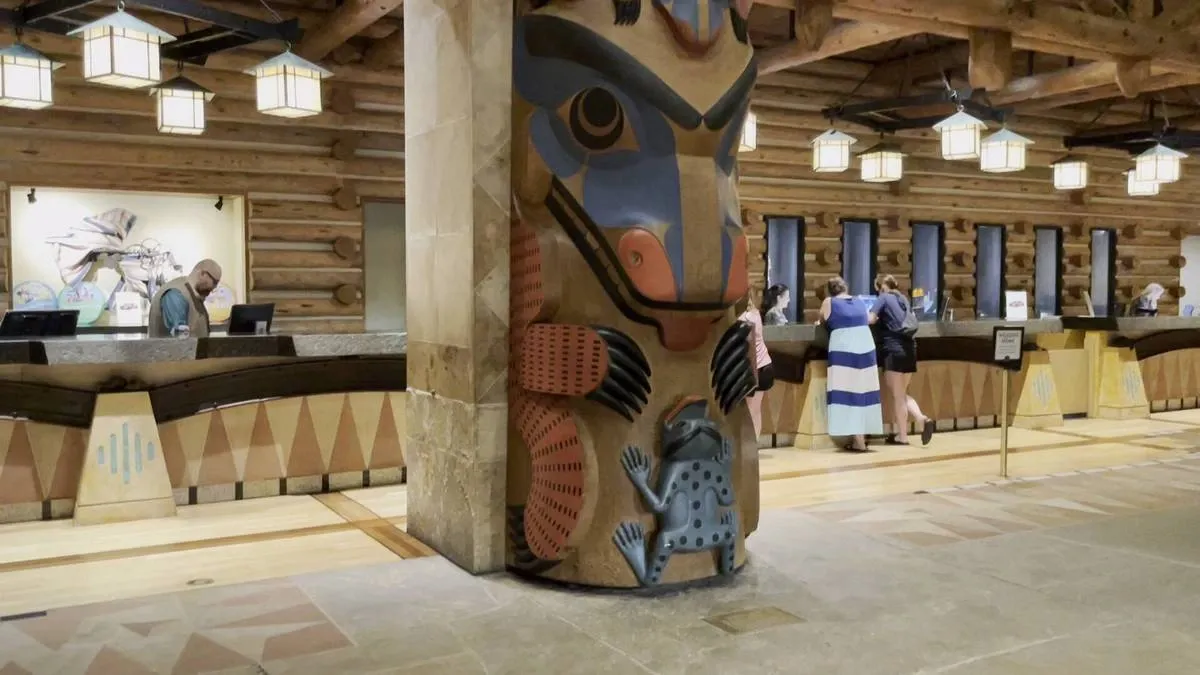 A hotel lobby features a rustic wooden reception desk with a staff member assisting guests. A prominent totem pole with colorful, intricate designs stands in the foreground. 