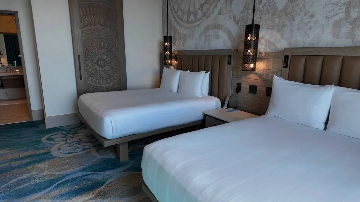 A hotel room features two neatly made double beds with white linens and padded headboards. Decorative pendant lights hang above the bedside table, and the room has a blue patterned carpet and a stylish mural on the wall.