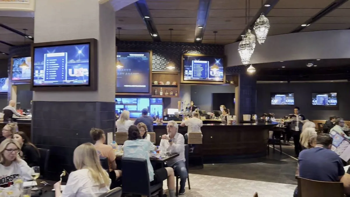 A restaurant scene with people enjoying their meals. Multiple large screens display sports content, and a bar area is visible in the background.