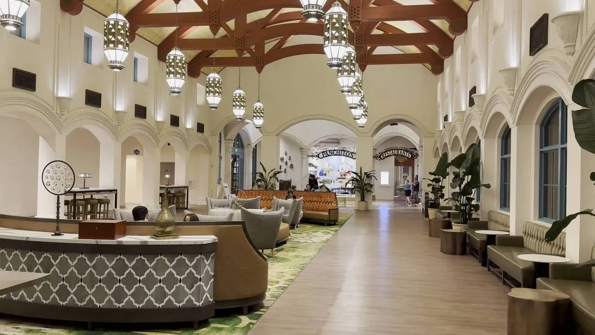 A spacious hotel lobby with a high ceiling featuring wooden beams and elegant hanging lanterns. The area is decorated with plush seating, patterned carpets, and potted plants.