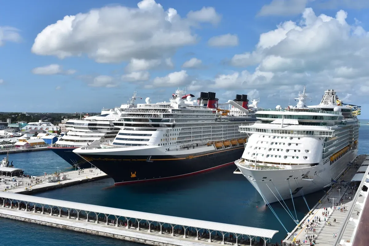 Several large cruise ships are docked at a bustling port under a partly cloudy sky, with passengers visible on the pier and vibrant buildings in the background.