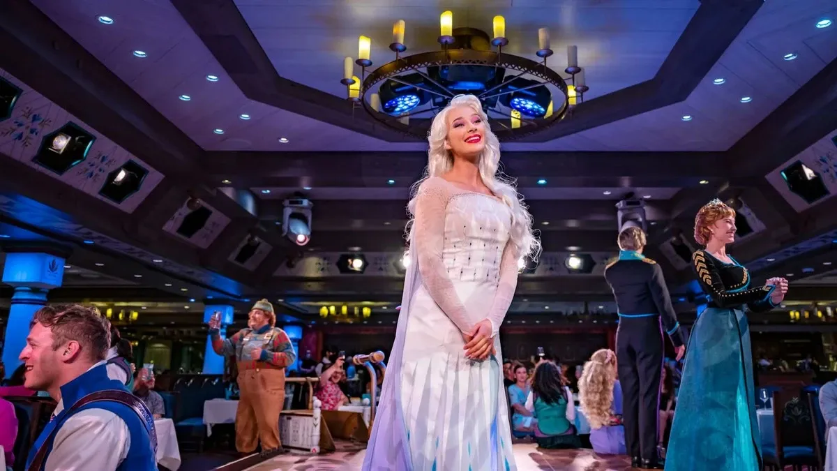 A performer dressed as a character in a white gown with long, white hair stands on a stage in a lively, colorful dining area, surrounded by other performers in themed costumes, while guests watch and enjoy the show.