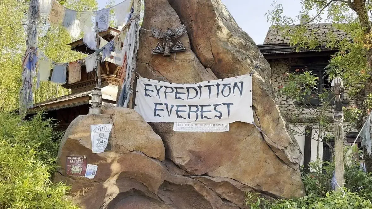 A large rock displays a sign reading "Expedition Everest" in a theme park setting. Colorful prayer flags and a rustic building with wooden details are visible in the background, surrounded by lush greenery.