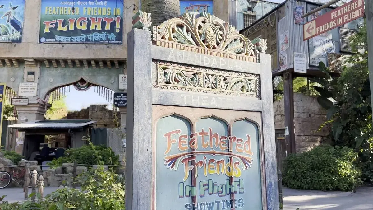 A decorative sign for "Feathered Friends in Flight" is prominently displayed, featuring colorful lettering and intricate carvings. In the background, there are additional signs and greenery, creating an inviting atmosphere for the show at Anandapur Theater.
