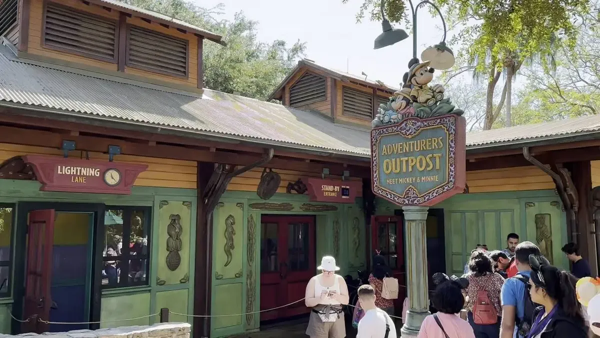 A colorful building with a corrugated metal roof features signs for "Adventurers Outpost" and "Lightning Lane." A crowd of people, some wearing Mickey Mouse ears, gather outside under a clear sky, surrounded by trees.
