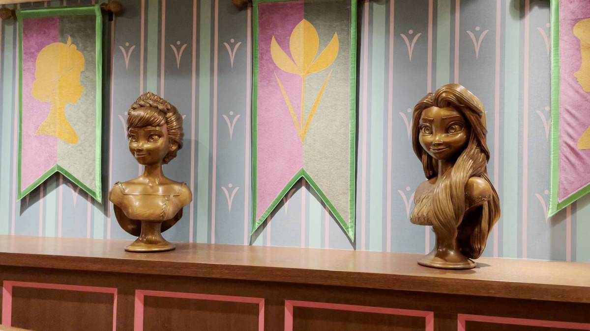 Two bronze busts of animated characters are displayed on a wooden shelf, set against a backdrop of pastel banners with floral designs and silhouettes.