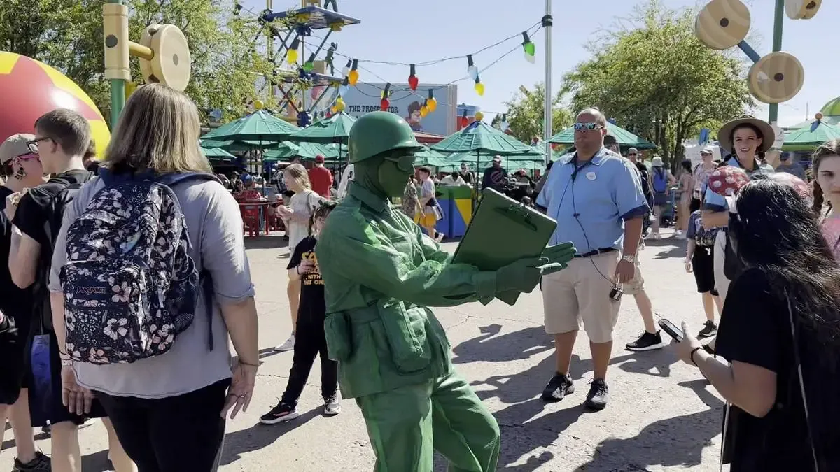 A person dressed as a green toy soldier interacts with visitors at Disney World, holding a clipboard. There's people gathered around, and colorful decorations like oversized toys and string lights in the background.