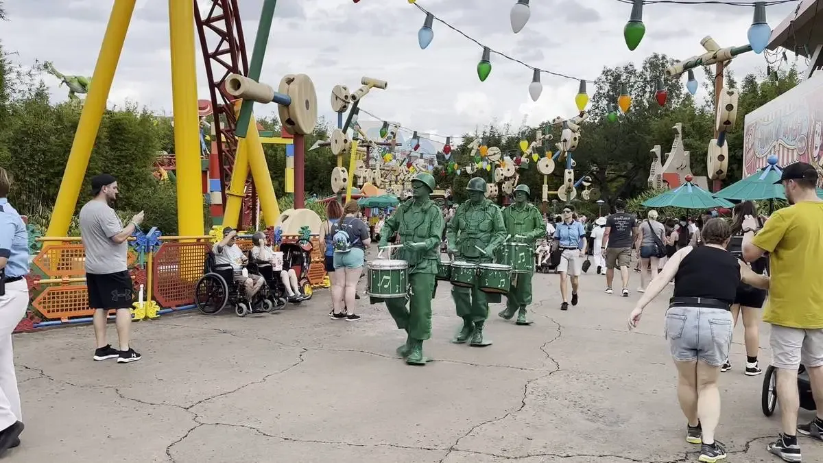 Three performers dressed as green toy soldiers march with drums in a lively theme park area, surrounded by colorful oversized toy decorations and bustling visitors.