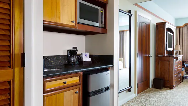 A cozy hotel room kitchenette features wooden cabinets, a microwave, a small sink, and a mini-fridge. A coffee maker sits on the black countertop, and a large mirror is mounted on the wall. The room extends to a living area with a TV and wooden furniture.