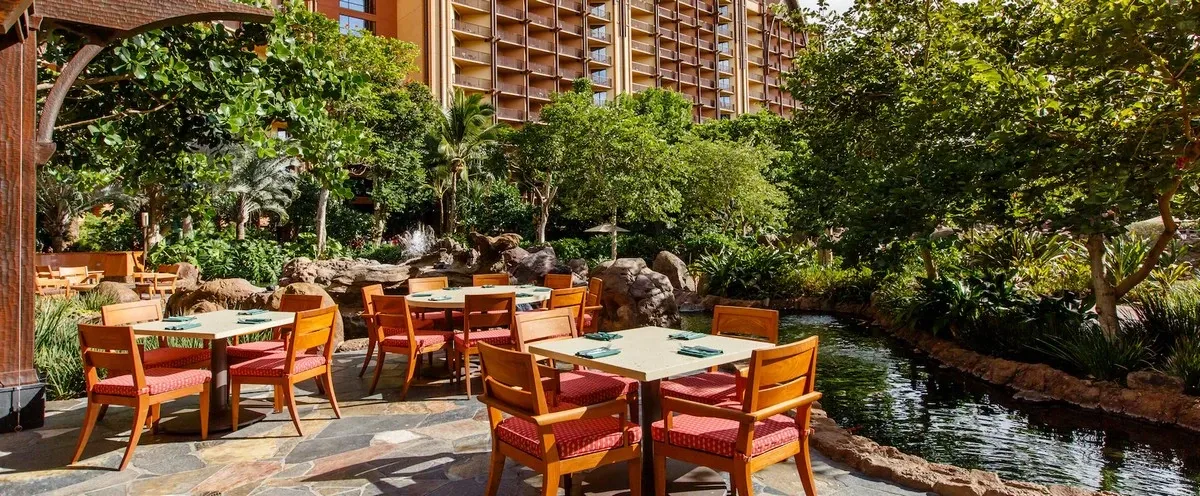 A charming outdoor dining area features several tables with red-cushioned wooden chairs, set on a stone patio beside a small, tranquil stream. The lush greenery and towering hotel building in the background create a serene, tropical atmosphere.