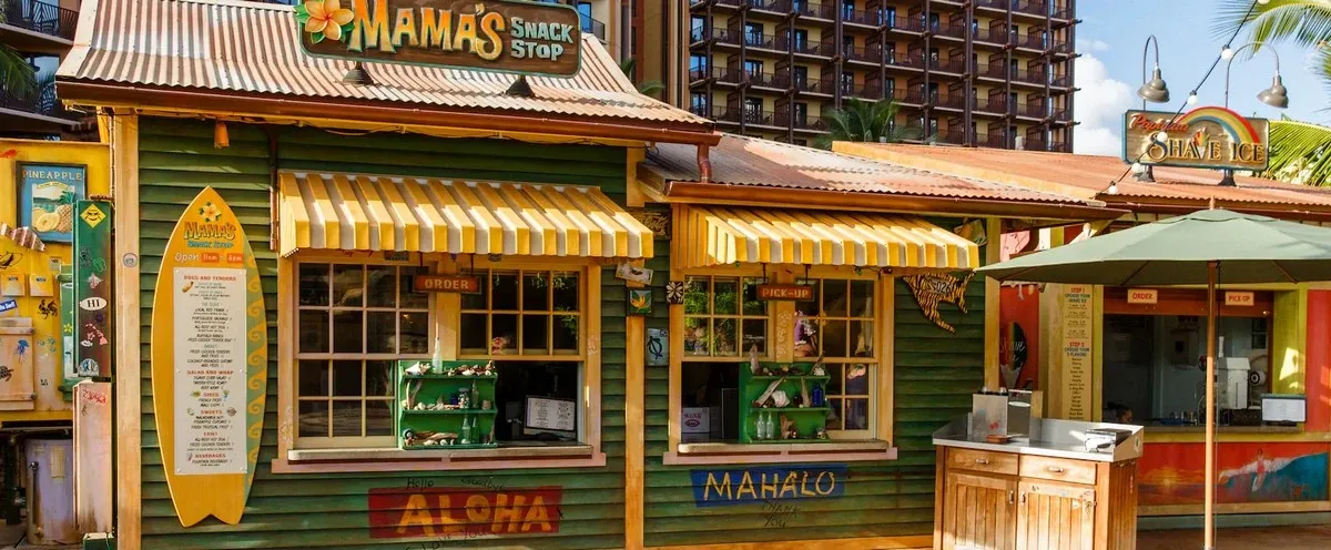 A colorful snack stand with a corrugated metal roof, featuring "Mama's Snack Stop" signage and a surfboard menu. The stand has yellow and white striped awnings, with open service windows displaying snacks. A nearby shave ice booth and palm trees add to the tropical setting.