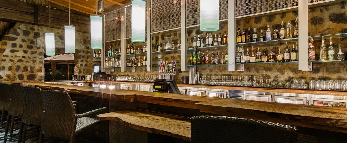 A cozy bar interior featuring a long wooden counter with wicker chairs, illuminated by soft pendant lights. The stone wall in the background showcases a variety of liquor bottles neatly arranged on shelves, creating an inviting atmosphere.