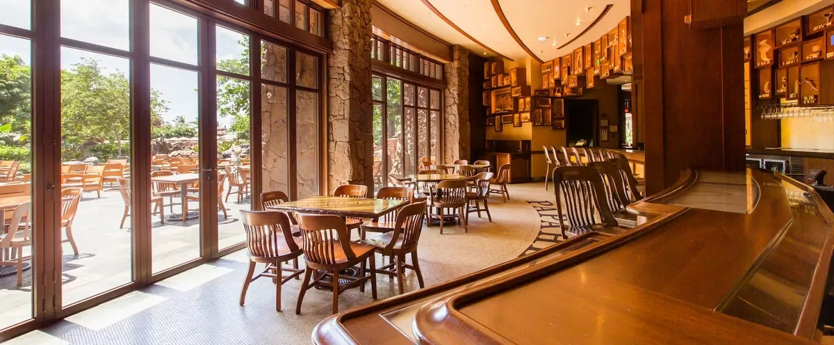 A cozy restaurant interior with wooden tables and chairs, featuring large windows that open to a sunlit outdoor patio with more seating. The warm wooden tones and soft lighting create an inviting atmosphere, while framed pictures adorn the walls, adding character to the space.