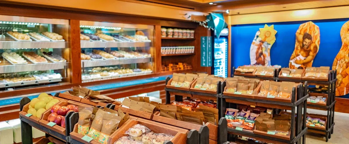A cozy bakery features wooden shelves filled with various baked goods in brown paper bags, alongside a display of fresh apples and other fruits. In the background, glass cases showcase an assortment of pastries and breads, while vibrant artwork decorates the walls, adding a colorful touch to the warm setting.