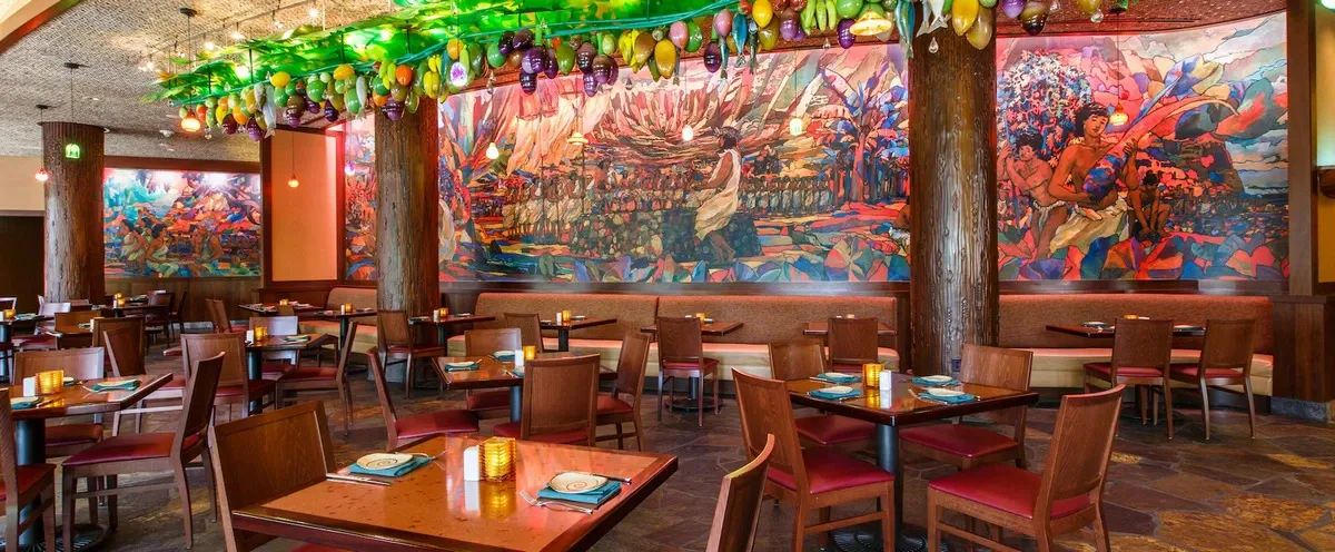 A vibrant restaurant interior featuring colorful murals on the walls, depicting lively scenes. The ceiling is adorned with hanging fruit decorations, and the room is filled with wooden tables and chairs set with plates and napkins, creating a warm and inviting atmosphere.