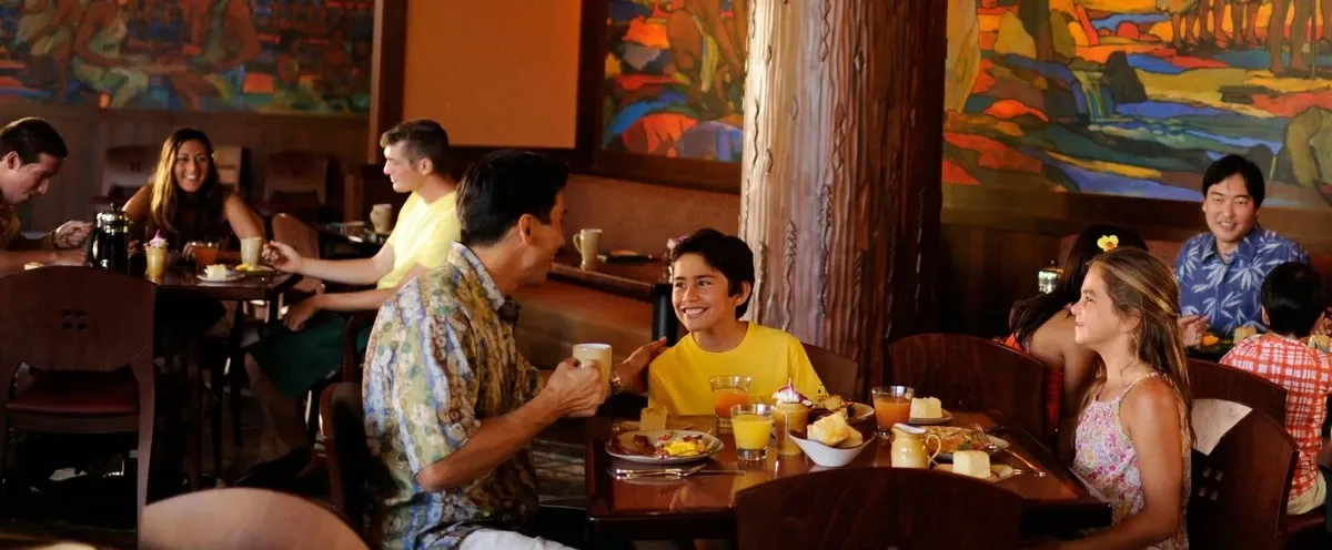 A group of people is enjoying a meal in a warmly lit restaurant with colorful murals on the walls. A man in a patterned shirt converses with two children at a table set with breakfast items, including juice and bread. Other diners are visible in the background, creating a lively atmosphere.
