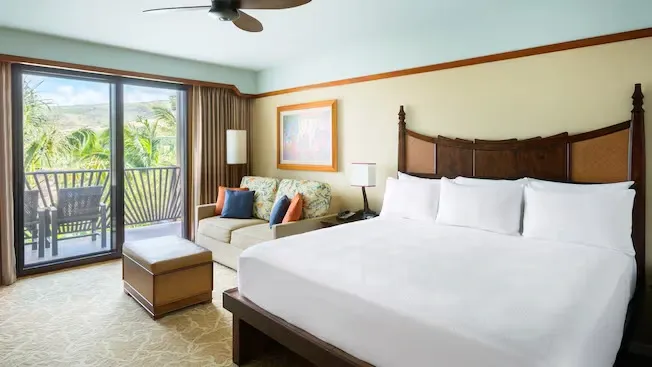 A bright hotel room features a large bed with white linens, a wooden headboard, and a cozy sofa with colorful cushions. A glass door opens to a balcony with a view of lush greenery, and a ceiling fan hangs above.