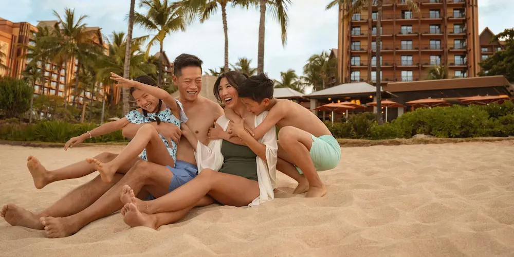 A family of four sits joyfully on a sandy beach, laughing together. Behind them, palm trees and a resort building are visible. The parents are in swimwear, with their children playfully leaning on them, creating a warm and happy scene.