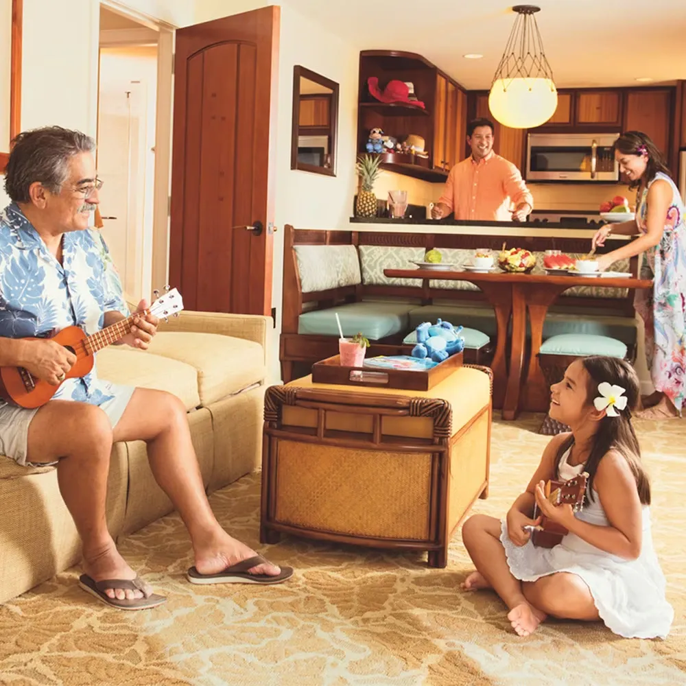 A man sits on a couch playing a ukulele, while a young girl with a flower in her hair sits on the floor, also holding a ukulele, smiling at him. In the background, a man and woman are preparing food in a cozy kitchen with wooden cabinets and a warm atmosphere.