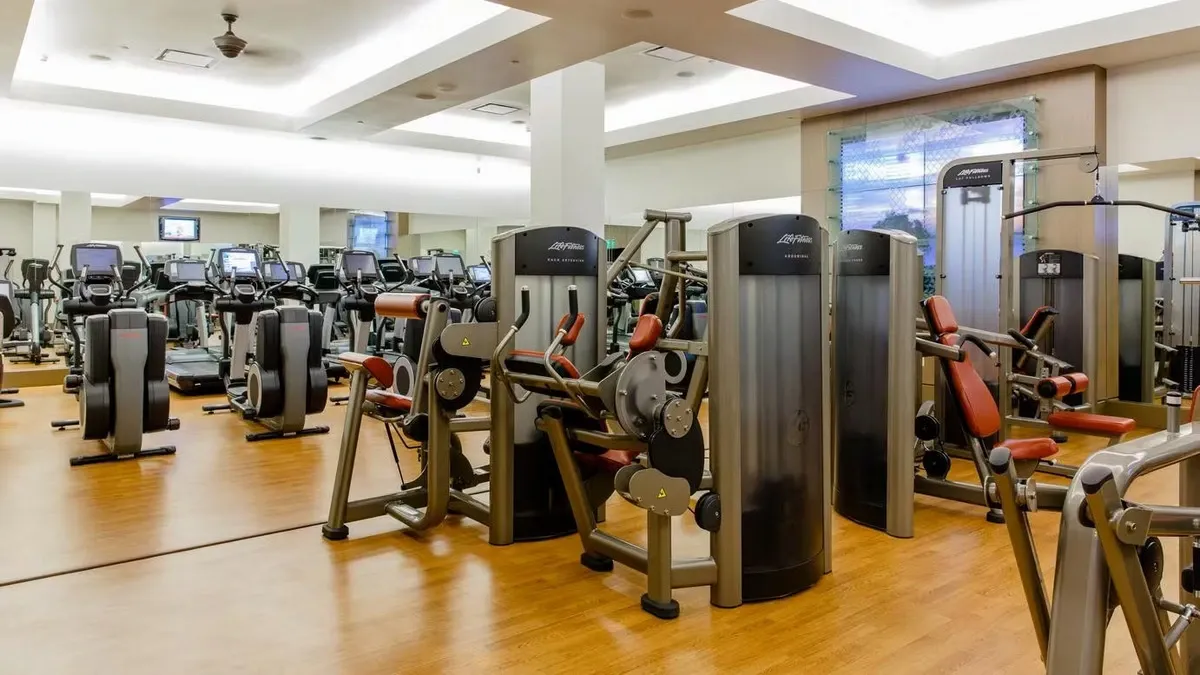 A spacious gym with a variety of exercise machines, including ellipticals and strength-training equipment, set on a polished wooden floor with large mirrors reflecting the bright, well-lit interior.