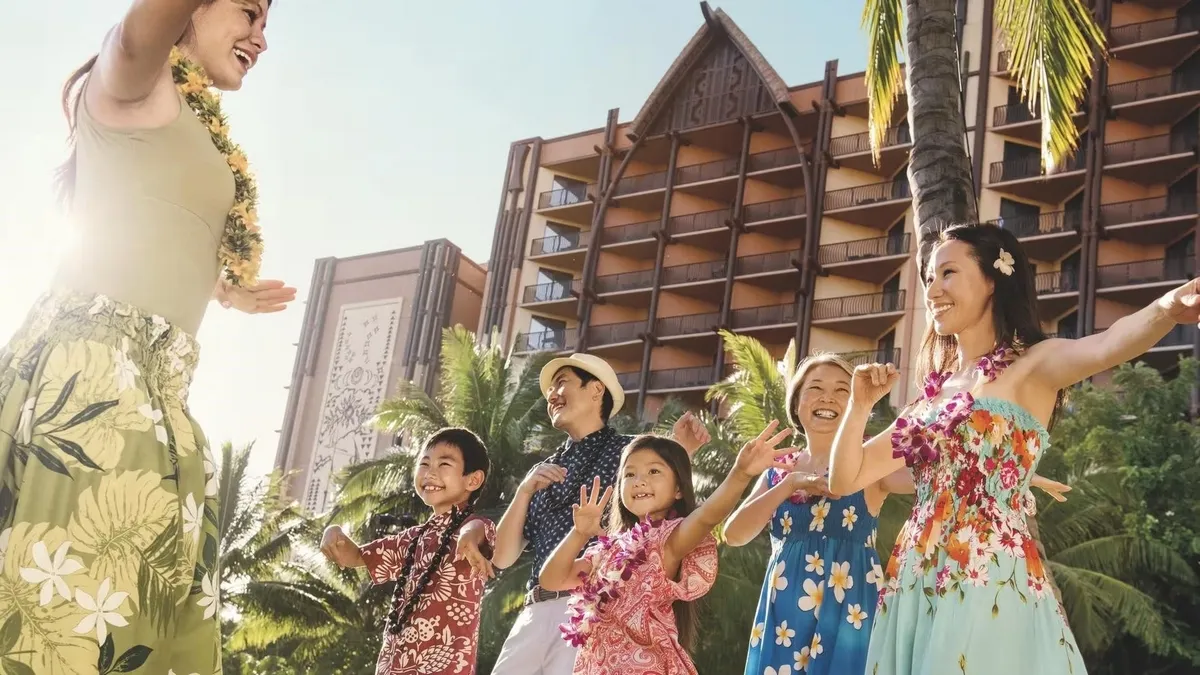 A group of people, including adults and children, are joyfully learning a hula dance from an instructor in front of a tropical resort. They are wearing colorful floral clothing and leis, surrounded by palm trees and the sunlit facade of the hotel.