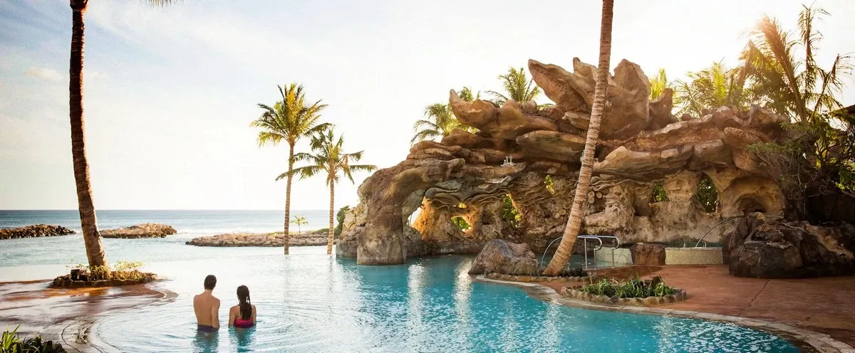 A couple stands in a tranquil pool, gazing at the ocean, surrounded by palm trees and a rocky grotto under a clear sky.