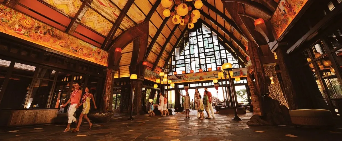 A group of people walks through a spacious, warmly lit lobby with high wooden ceilings and intricate wall decorations. Large windows and hanging lamps add to the inviting atmosphere.