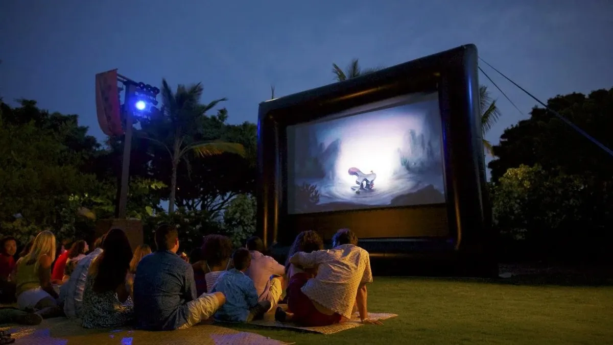 A group of people sits on a grassy lawn watching a movie projected on a large inflatable screen under the evening sky, surrounded by palm trees and soft lighting.