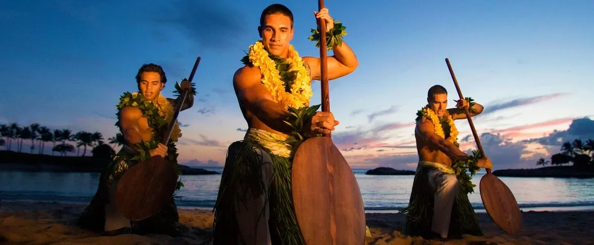 Three men dressed in traditional Polynesian attire perform a dance with wooden paddles on a beach at sunset, adorned with leafy garlands and leis, with a serene ocean and silhouetted palm trees in the background.