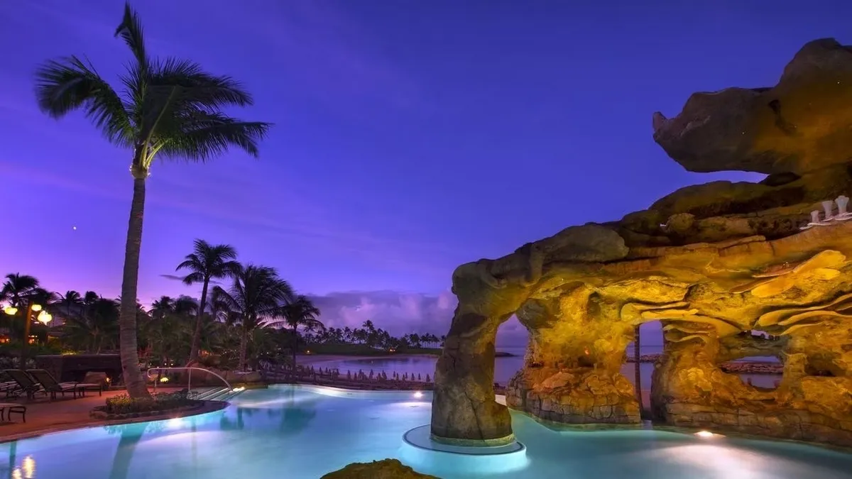 A serene poolside scene at dusk features a large rock formation partially overhanging a glowing pool, with palm trees silhouetted against a vibrant purple sky.