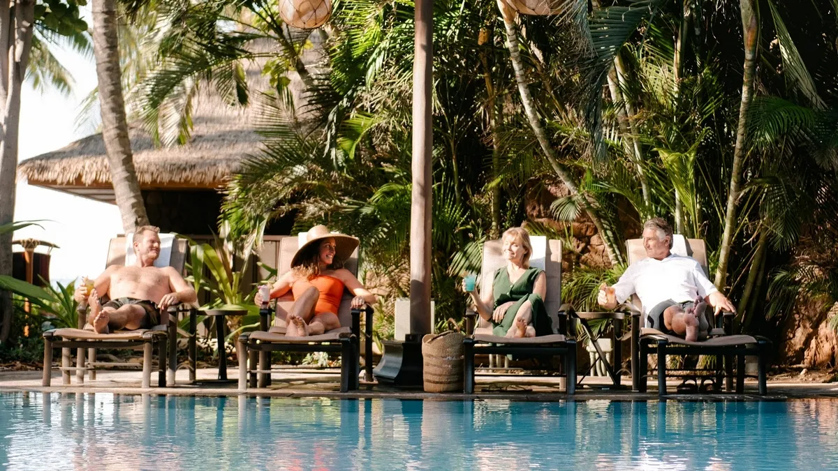 Four people relax on lounge chairs by a pool, enjoying drinks under the shade of palm trees, with a thatched-roof hut in the background.