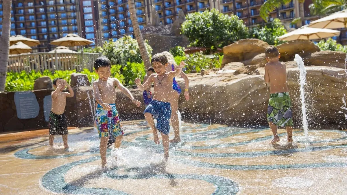 Children joyfully play in a splash pad with water jets, surrounded by lush greenery and resort buildings in the background.