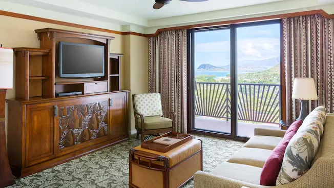 A cozy living room features a wooden entertainment center with a TV, a patterned armchair, and a sofa with floral cushions. The room opens to a balcony with a scenic view of lush greenery and distant mountains through large glass doors.