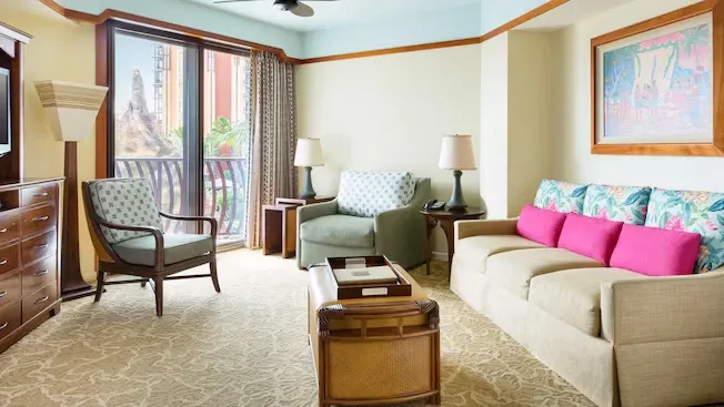 A cozy living room features a beige sofa with bright pink and patterned cushions, a matching armchair, and a wooden coffee table. Large windows with sheer curtains offer a view of lush greenery, while a colorful framed artwork adorns the wall. The carpet has a subtle, decorative pattern.