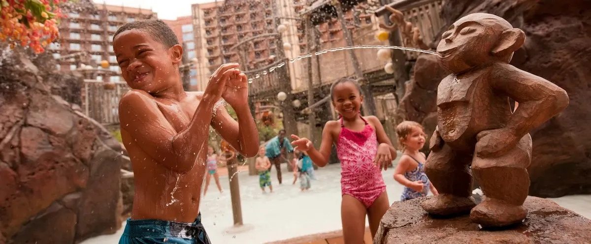 Children play joyfully in a water area, with a boy in blue swim trunks laughing as he gets splashed by a monkey-shaped fountain. A girl in a pink swimsuit smiles nearby, while other children enjoy the background, surrounded by rocky structures and a resort-like setting.